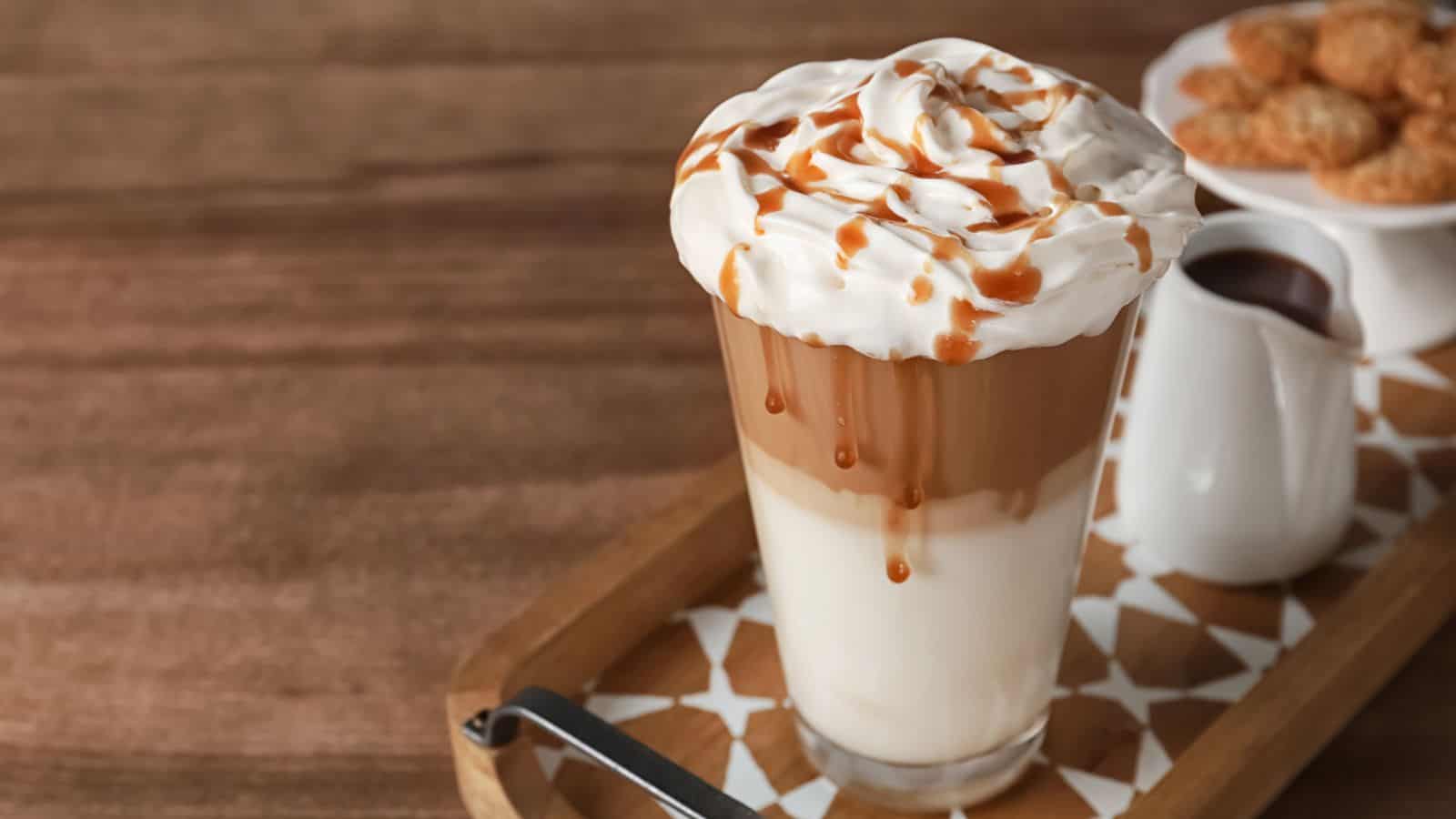 A glass of layered iced coffee topped with whipped cream and caramel drizzle sits on a wooden tray. A small white pitcher and a bowl of cookies are in the background on a wooden table.