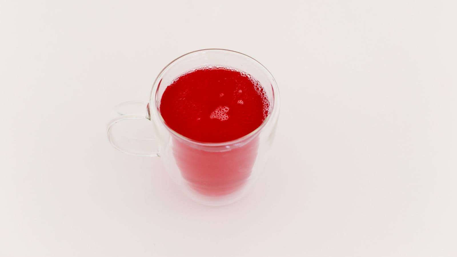 A clear glass mug filled with bright red liquid sits on a white surface. The mug has a handle and the contents appear to be juice or a similar beverage, with small bubbles visible on the surface.