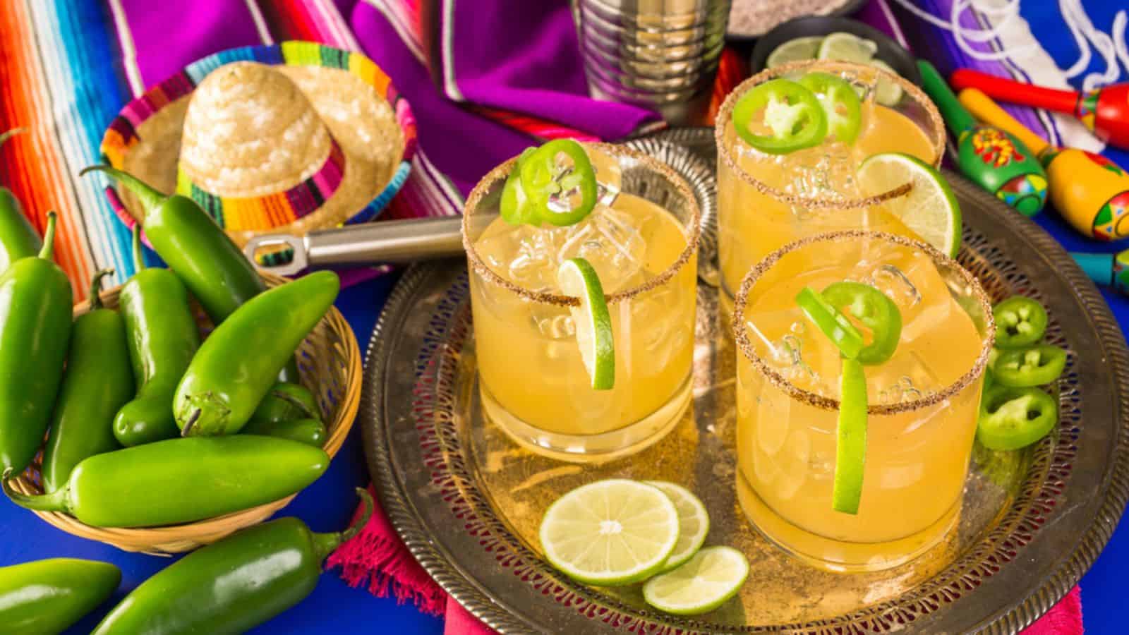Three glasses of orange cocktails with ice and jalape&ntilde;o slices on a metal tray. Nearby are sliced limes, whole jalape&ntilde;os, a colorful cloth, maracas, a basket of chips, and a cocktail shaker, set against a vibrant blue background.