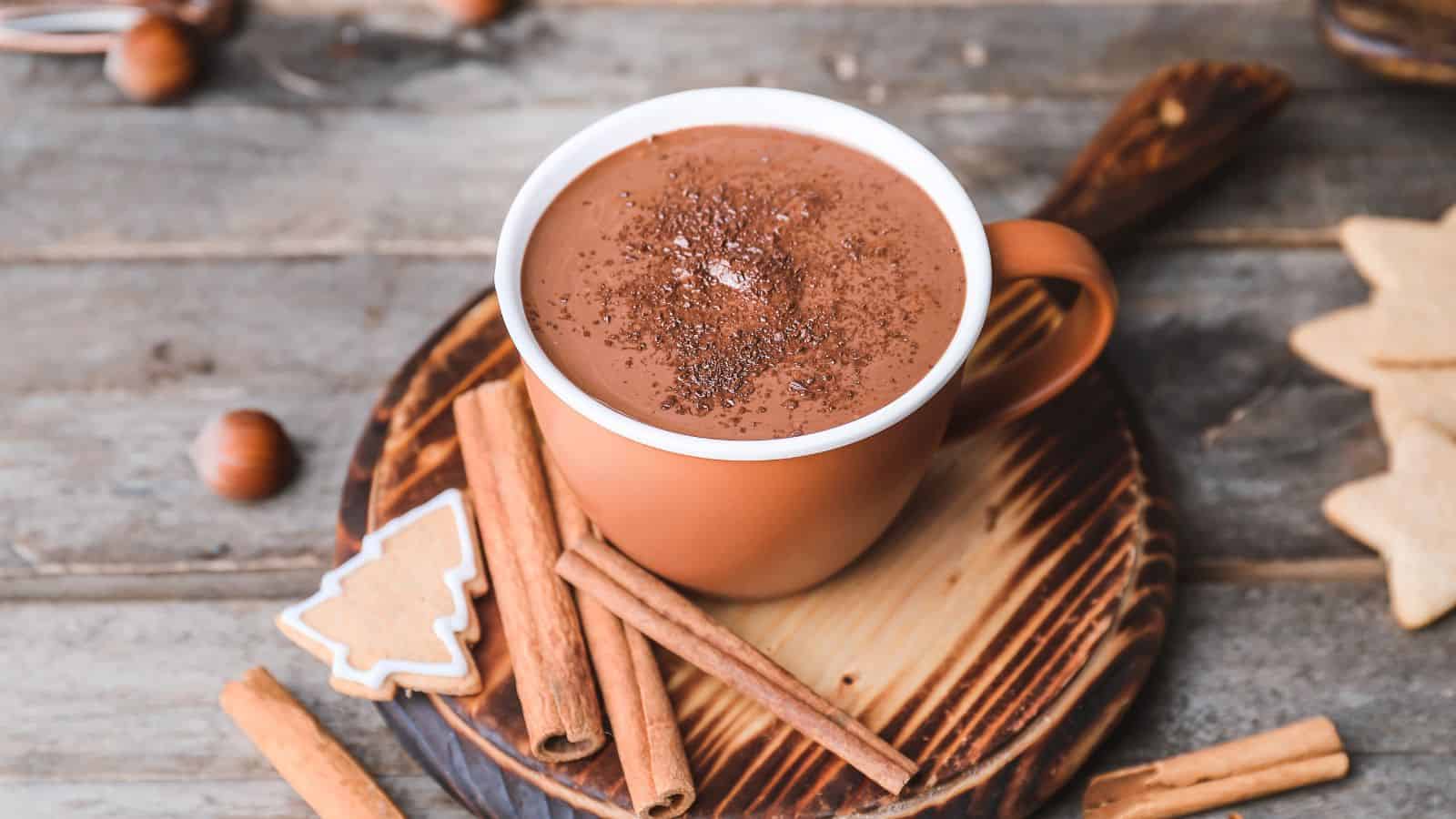 A ceramic mug filled with hot chocolate is placed on a wooden coaster. Cinnamon sticks and a tree-shaped iced cookie are next to the mug. The table is rustic wood, and hazelnuts are scattered around.