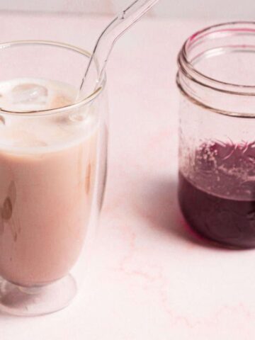 A glass of pink iced beverage with a clear straw next to a jar containing a dark purple liquid. Two cherries lie beside them on a light pink surface.