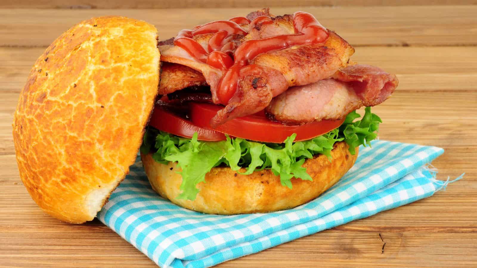A bacon and tomato sandwich with lettuce is placed on a round bread bun. The sandwich is topped with ketchup and rests on a light blue and white checkered cloth, on a wooden surface.