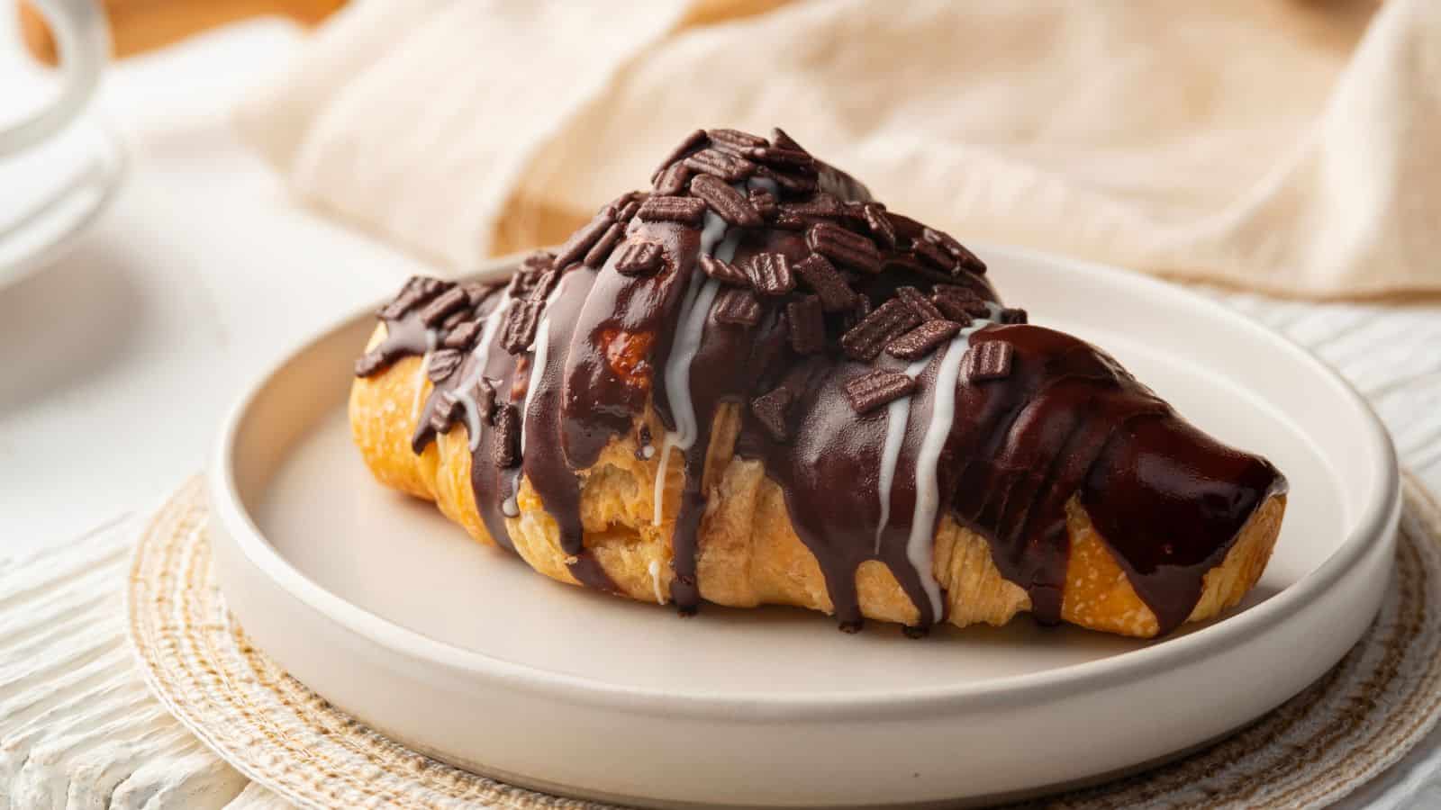 A croissant on a white plate is drizzled with chocolate and white icing and topped with small chocolate pieces. The plate rests on a woven placemat, and a beige cloth is partially visible in the background.