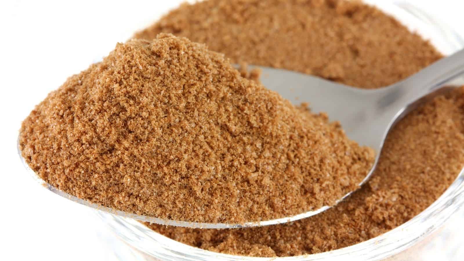 A close-up image shows a spoon filled with light brown ground cinnamon powder. The spoon hovers above a clear glass container also filled with the same powder. The background is white, highlighting the texture of the spice.