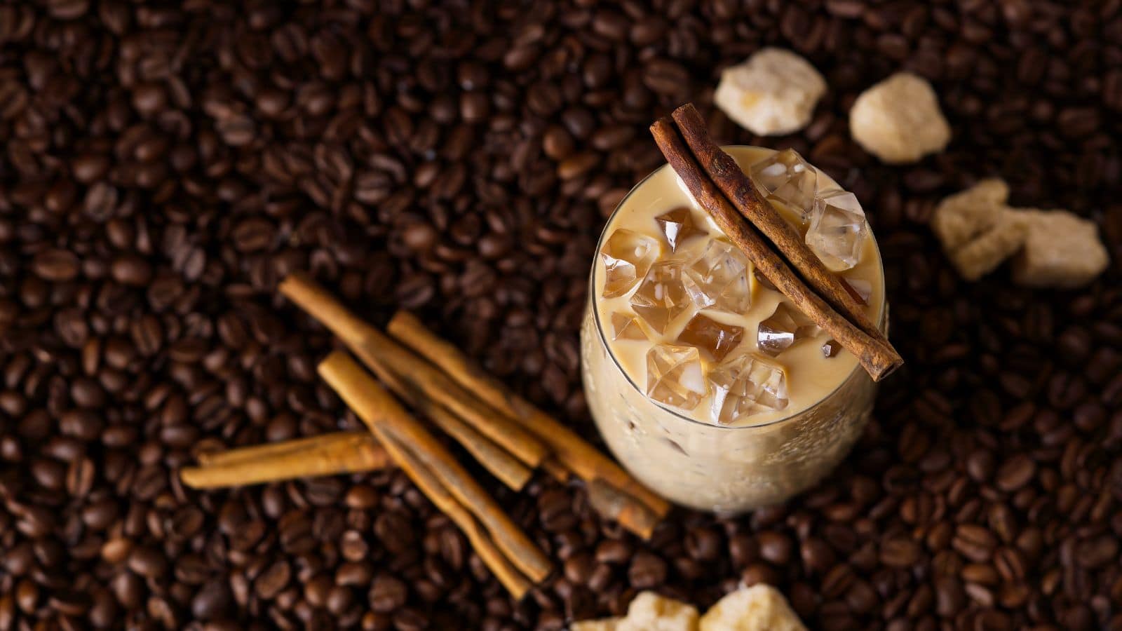 A glass of iced coffee with ice cubes and two cinnamon sticks on top is placed on a surface covered with coffee beans. Several whole cinnamon sticks and sugar cubes are scattered around the glass.