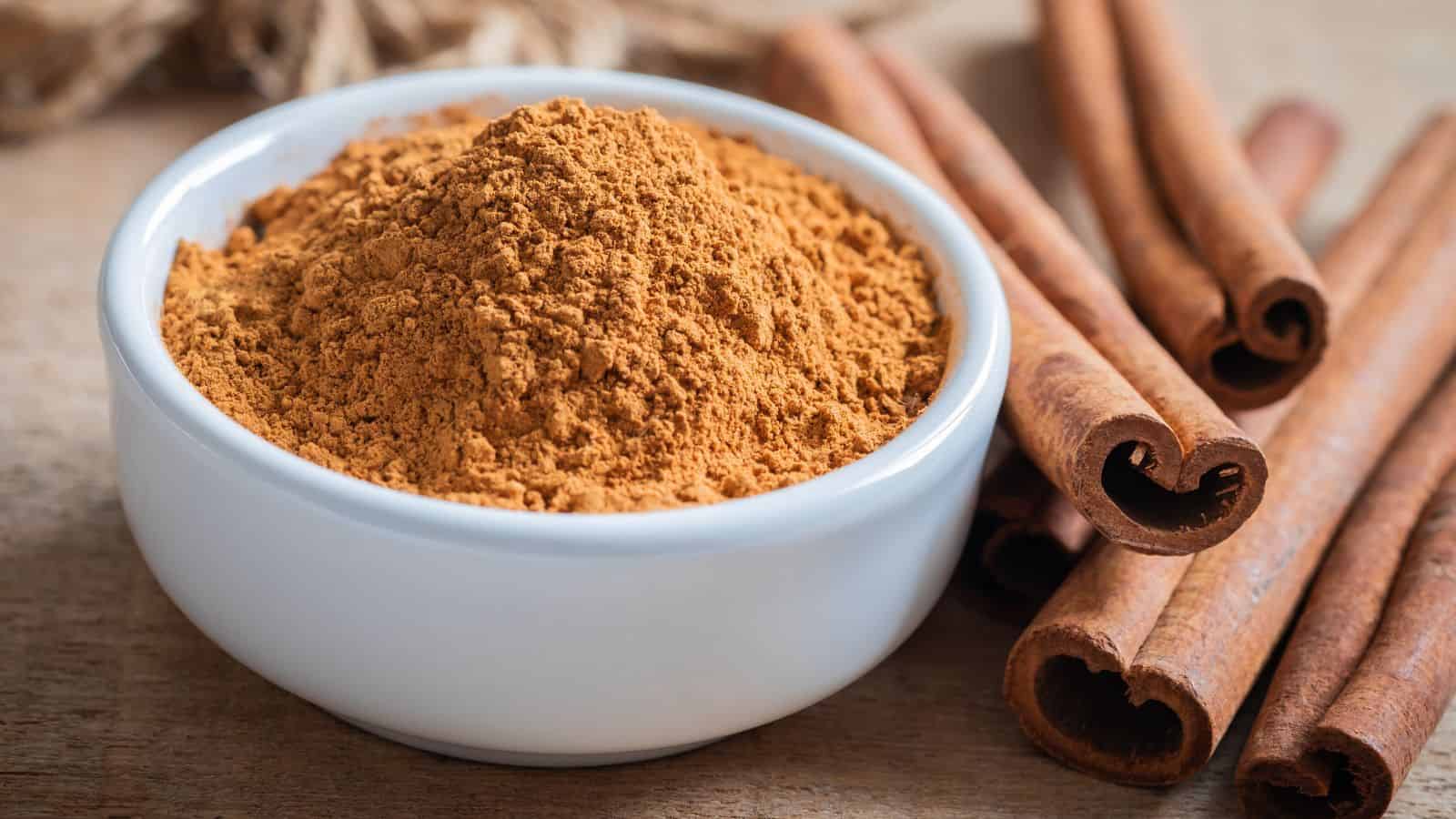 A white ceramic bowl filled with ground cinnamon is on a wooden surface. Several whole cinnamon sticks are placed to the right of the bowl, showcasing the spice in both its powdered and stick forms.