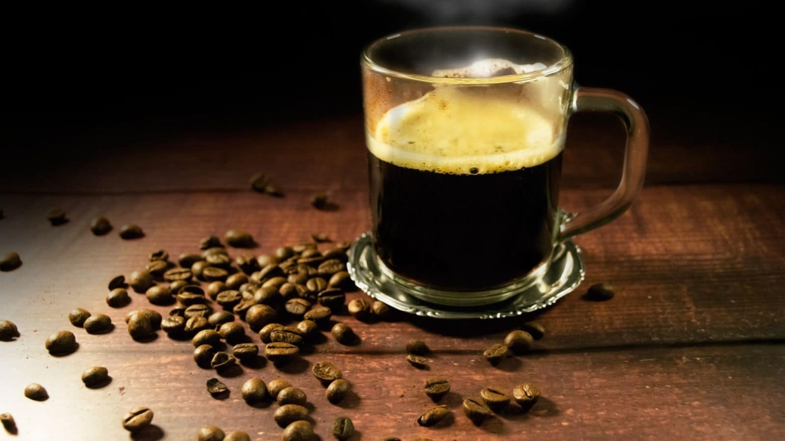 A glass cup filled with dark coffee sits on a wooden surface. Steam rises from the cup, indicating it's hot. Coffee beans are scattered around the cup, adding to the rustic ambiance. Lighting emphasizes the rich, dark color of the coffee.