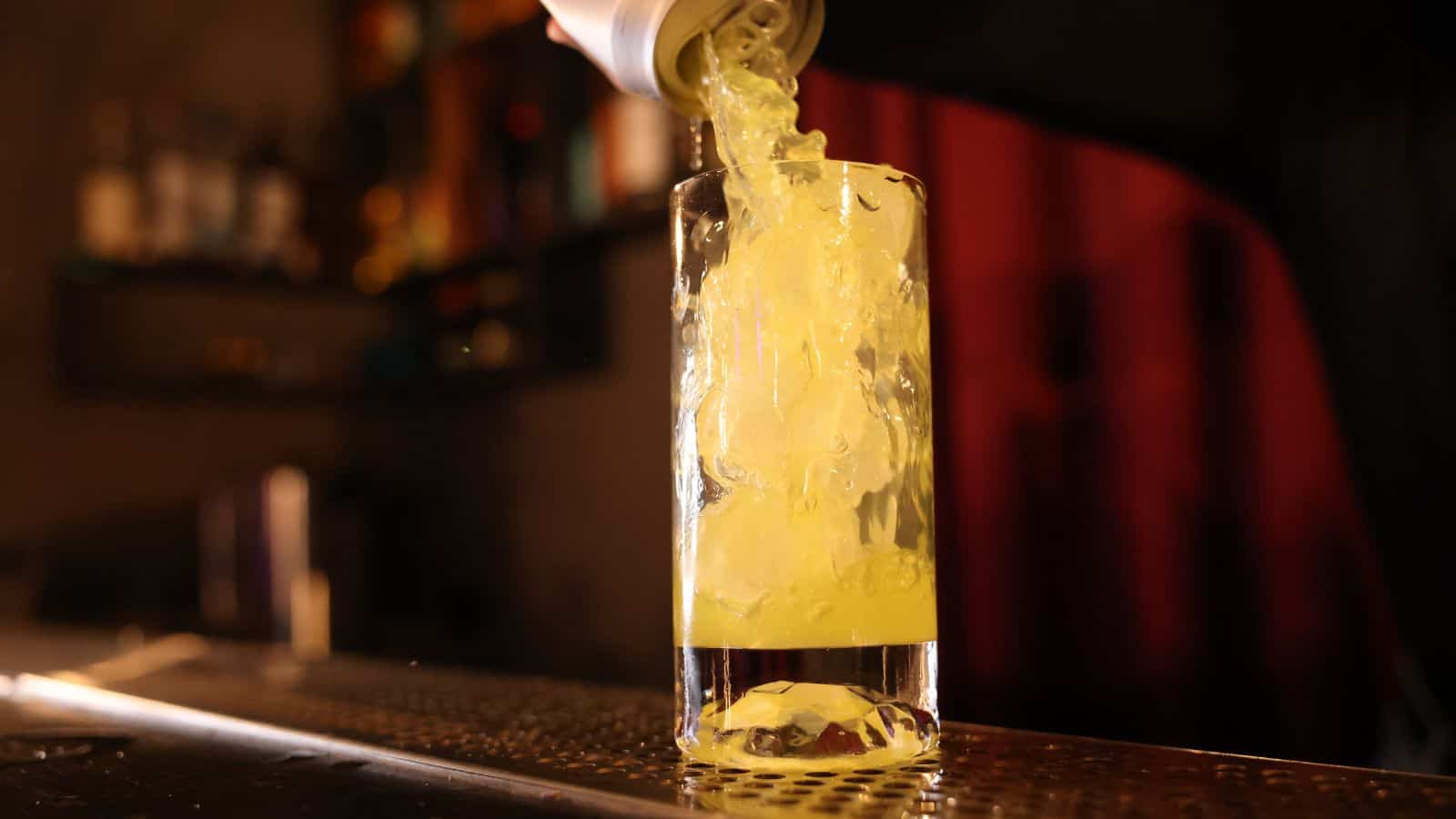 A can is pouring a yellow drink into a clear glass on a bar counter. The background is dimly lit with blurred shelves and bottles.