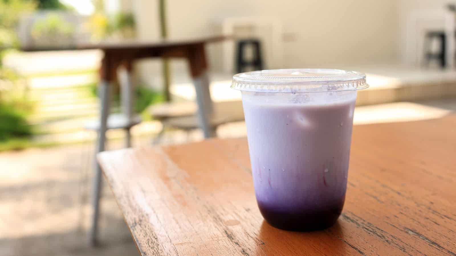 A plastic cup with a clear lid containing a purple and white layered drink sits on a wooden table. The background is a blurred outdoor seating area with chairs and tables. Natural light highlights the drink's gradient colors.