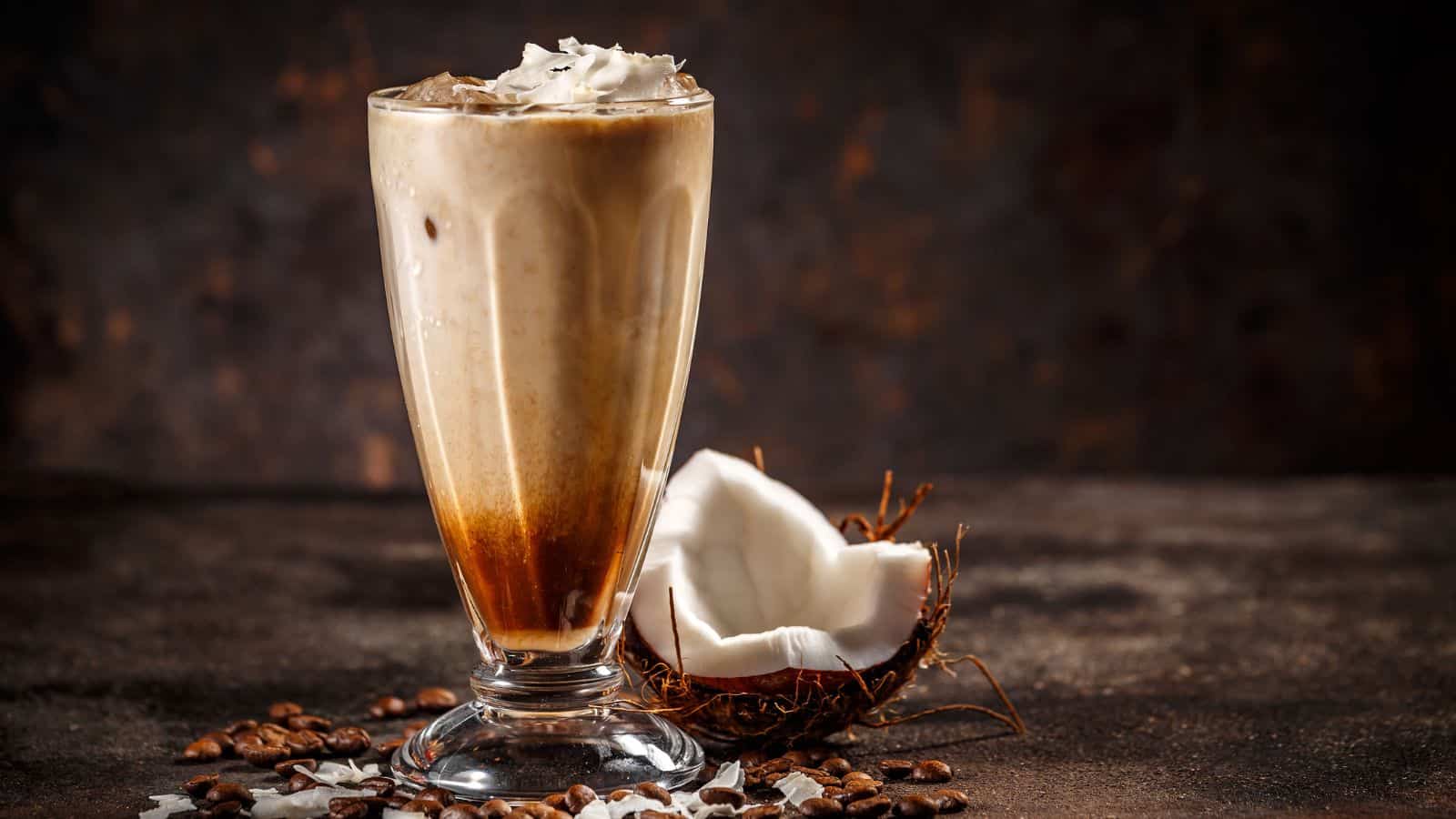 A tall glass of iced coffee topped with whipped cream is placed on a textured surface. Coffee beans and a half coconut shell are nearby, with coconut shavings scattered around. The background is dark and blurred.