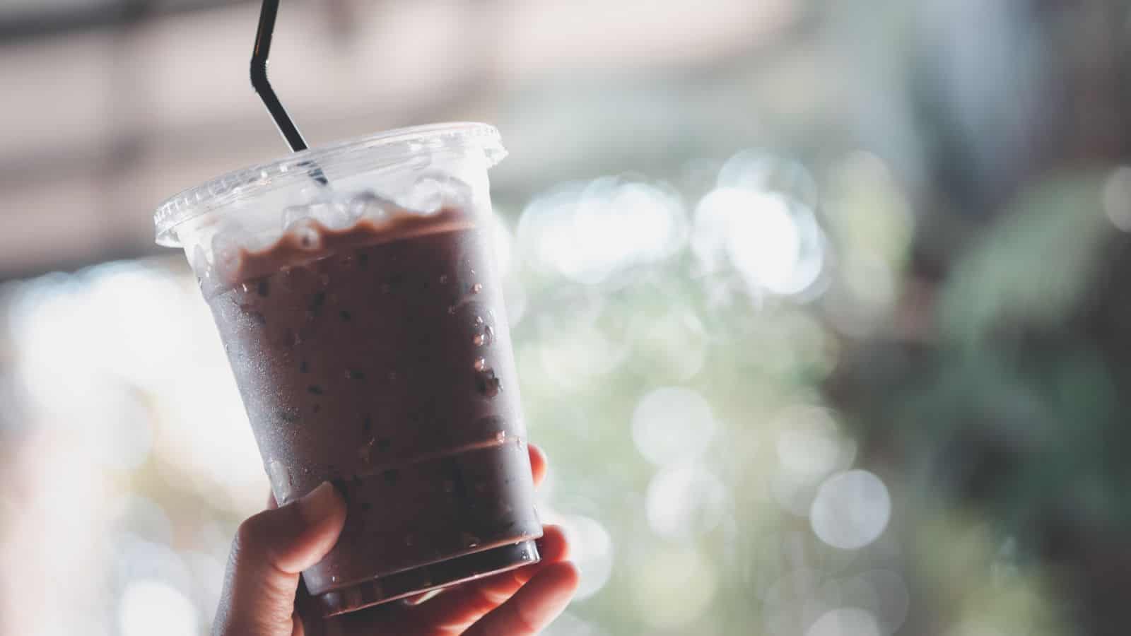 A hand holds a clear plastic cup filled with a cold chocolate beverage, topped with ice cubes. The cup has a black straw and a secure lid. The background is blurred with greenery and bokeh lights.