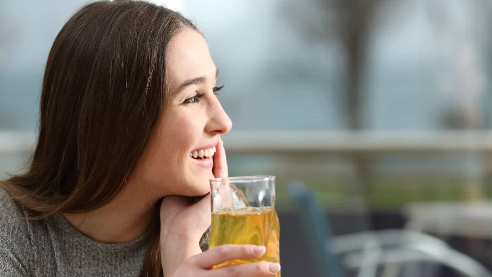 A person with long brown hair is smiling and holding a glass of yellow liquid. They are sitting outdoors with a blurred background of trees and sky.