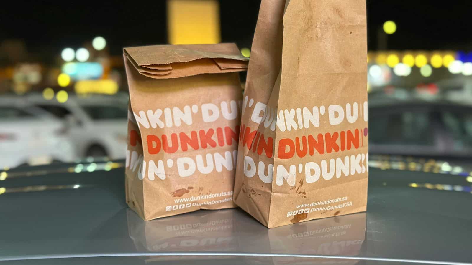 Two brown paper bags from Dunkin' are placed on a car roof. The bags display the brand's logo prominently. The background shows a blurred parking lot with cars and lights at night.