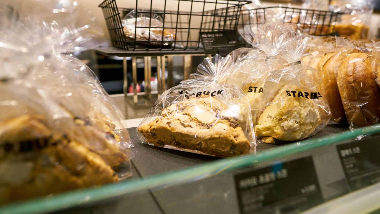 A display case showing various bakery items wrapped in clear plastic bags labeled with the Starbucks logo. Items include pie slices and bread, arranged in a neat line on a dark counter. A black wire basket is visible in the background.