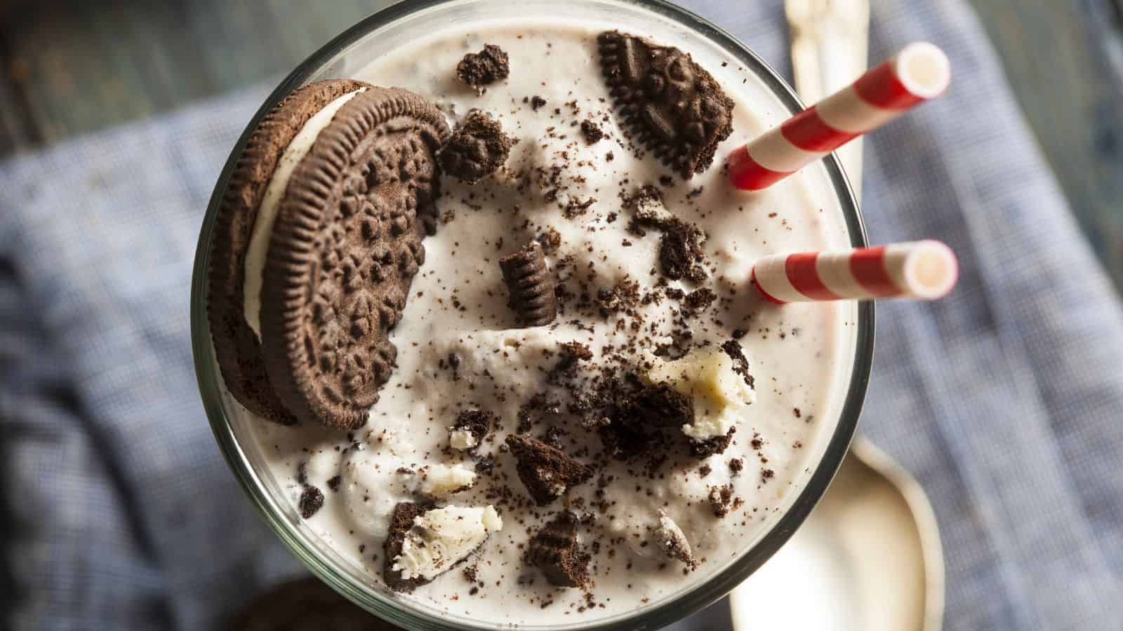 A glass filled with a cookies and cream milkshake is topped with crushed cookies. Two red and white striped straws are inserted into the drink. A whole cookie is placed on the rim. A spoon and a cloth are in the background.