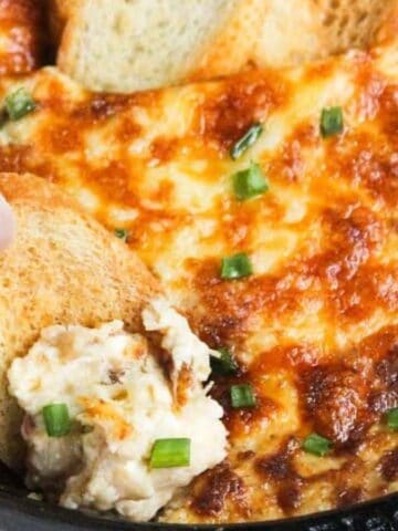 A hand dipping a slice of toasted bread into a cheese and herb dip that's baked until golden brown. The dish is garnished with chopped green onions and served in a black skillet.