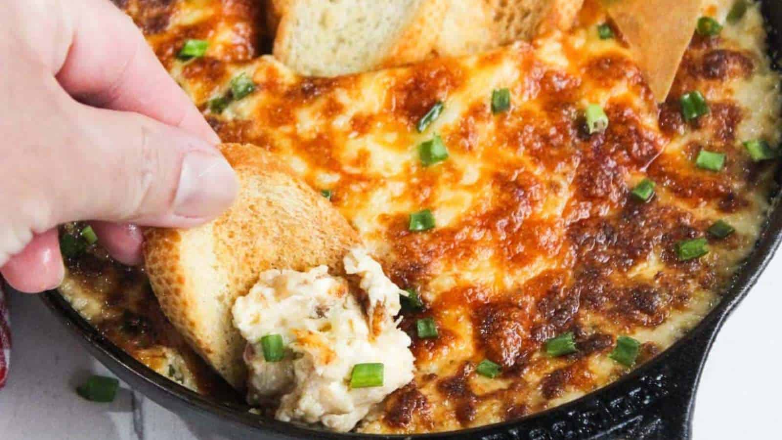 A hand dipping a slice of toasted bread into a cheese and herb dip that's baked until golden brown. The dish is garnished with chopped green onions and served in a black skillet.