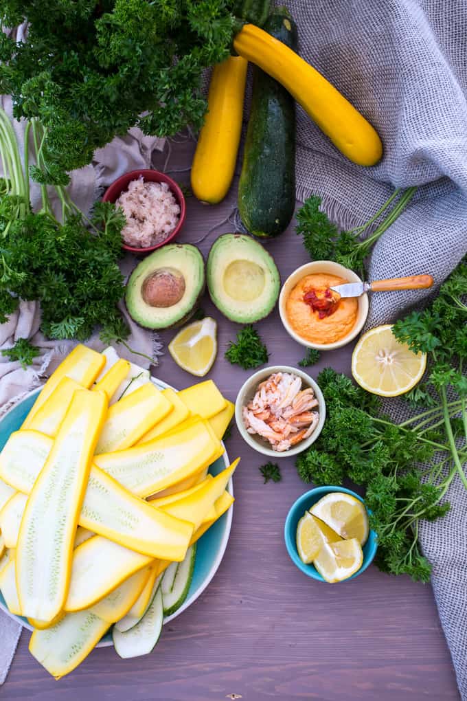 A wooden table showcases sliced yellow and green squash in a bowl, whole zucchinis perfect for zucchini roll up appetizers, halved avocado, parsley, lemon halves, and small bowls with salmon, hummus, and mysterious toppings. A grey cloth elegantly drapes part of the display.