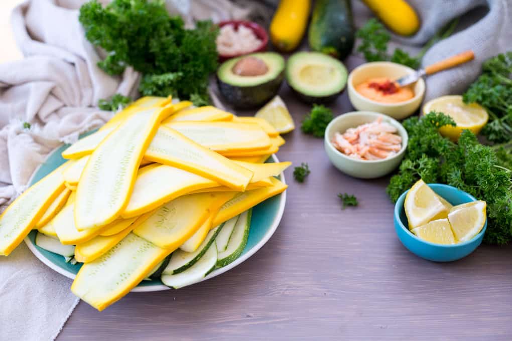A plate of thinly sliced yellow zucchinis, perfect for zucchini roll up appetizers, sits on a table surrounded by avocados, parsley, and lemon wedges. Small bowls hold various ingredients nearby, while a light cloth is draped elegantly beside them.