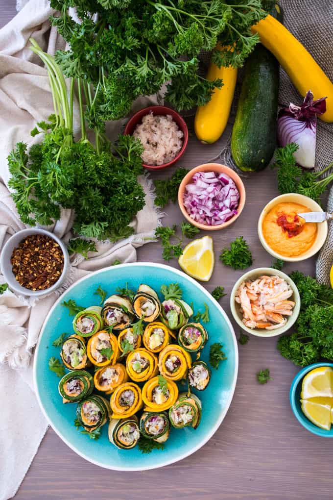 A platter of zucchini roll up appetizers, filled with stuffing, is surrounded by fresh parsley, lemons, yellow squash, zucchini, and small bowls of chopped onions, red pepper flakes, spreads, and a vegetable mix on a wooden table.