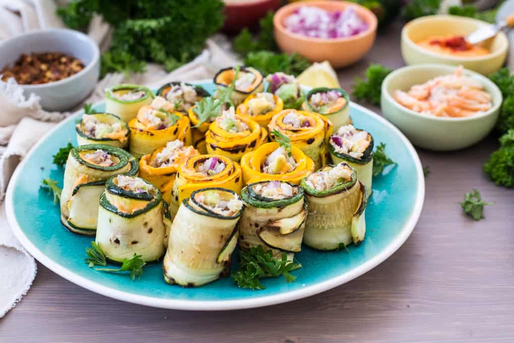 A plate of zucchini roll-up appetizers featuring rolled zucchini and yellow squash slices filled with a creamy mixture. The rolls are garnished with parsley and served on a blue plate, while various bowls with chopped onions, spices, and a creamy dip adorn the background.