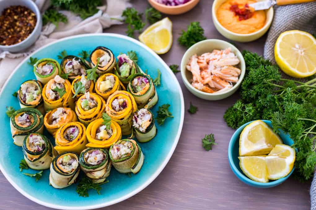 A plate of zucchini roll up appetizers features vegetable rolls made from zucchini and yellow squash, filled with a mix of chopped vegetables and herbs. Surrounding bowls contain almonds, orange sauce, and shredded chicken. Lemon wedges and fresh parsley complete the table setting.