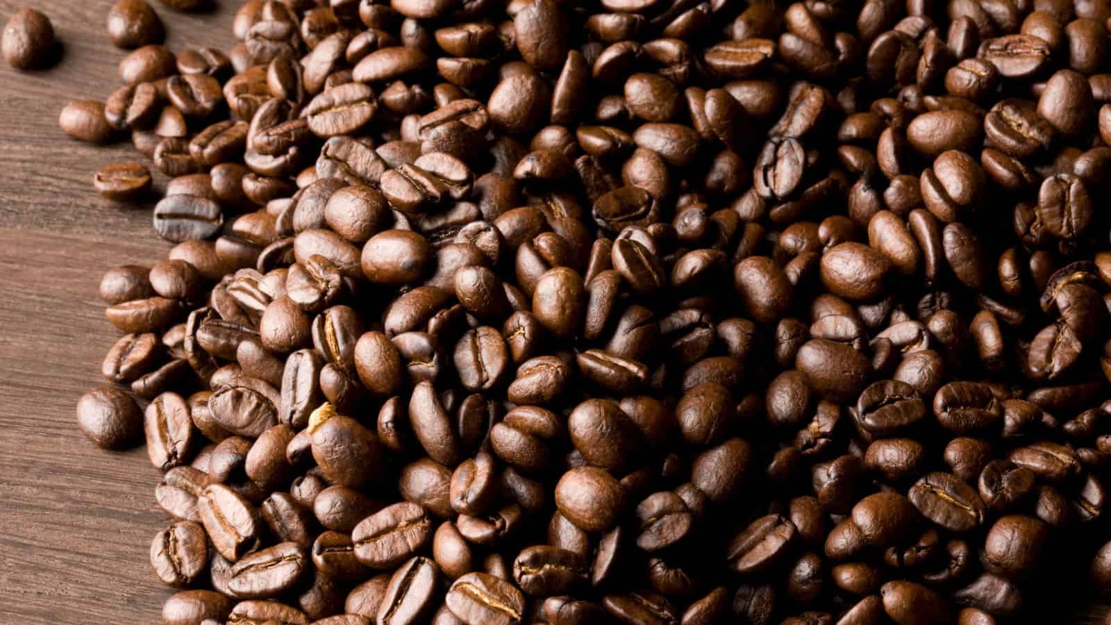A large pile of roasted coffee beans is spread across a wooden surface. The beans are glossy, with varying shades of dark brown, displaying a rich texture and an assortment of sizes.