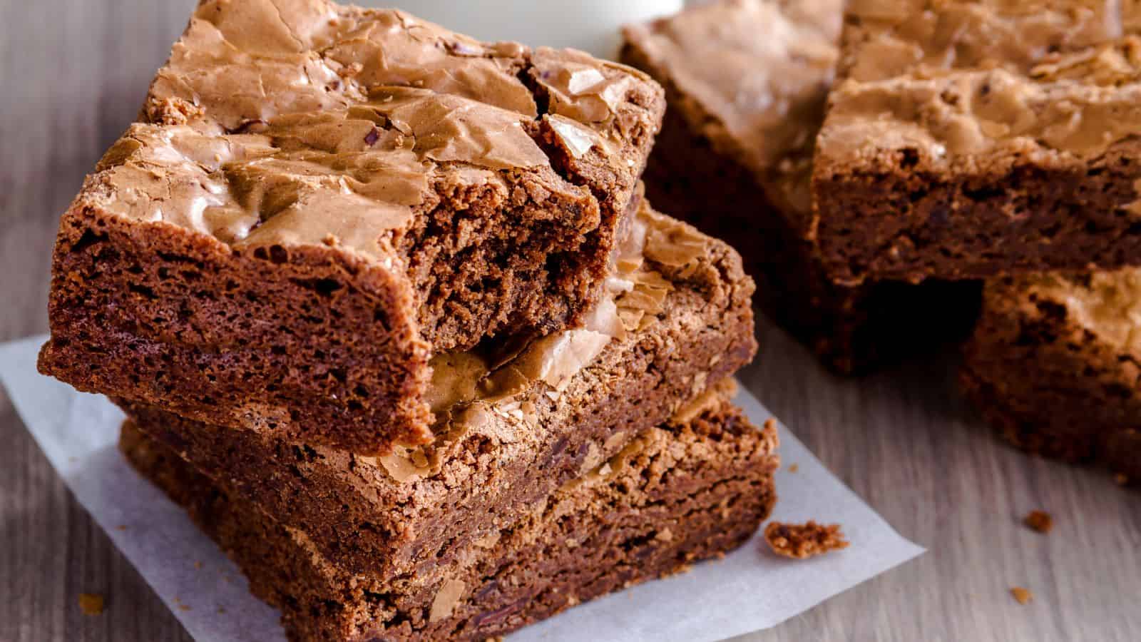 A stack of three chocolate brownies with a crackled top is placed on a piece of parchment paper. Some brownies are slightly overlapping in the background. The texture appears moist and fudgy, and they rest on a wooden surface.