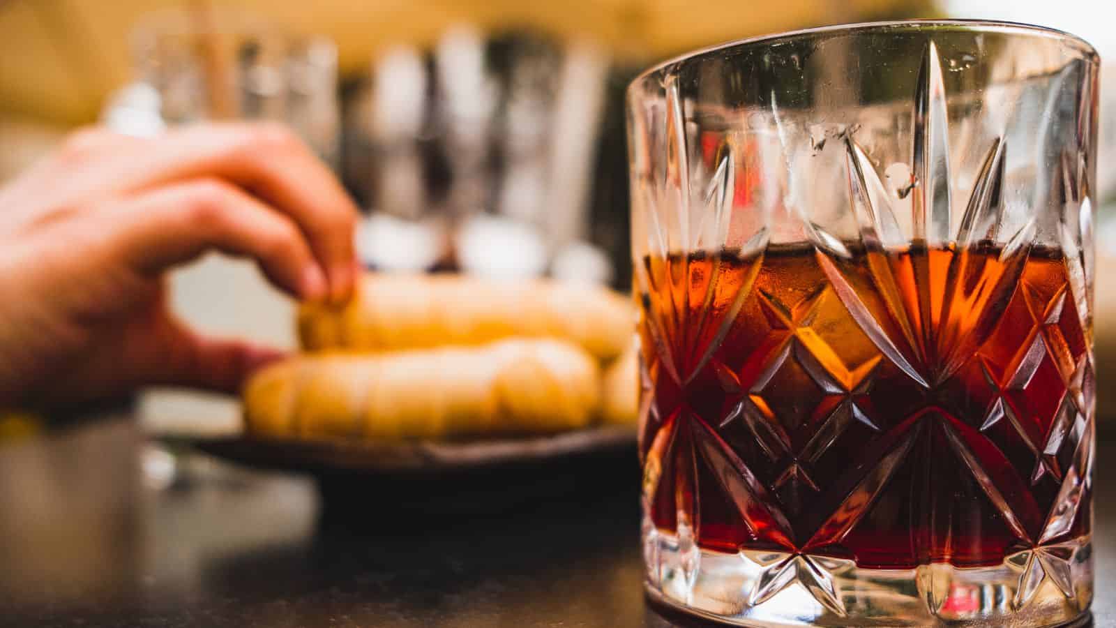 A glass with a dark-colored beverage sits on a table. The glass has a textured, geometric pattern. In the background, a hand reaches for a plate with croissants, partially out of focus.