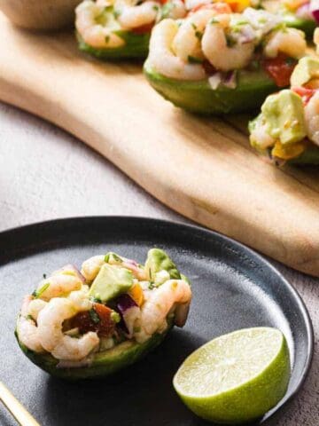A black plate with a halved avocado filled with a shrimp salad, garnished with lime. A fork rests beside it. A wooden board holds more avocado halves in the background. A wooden bowl with mixed salad ingredients is partially visible.