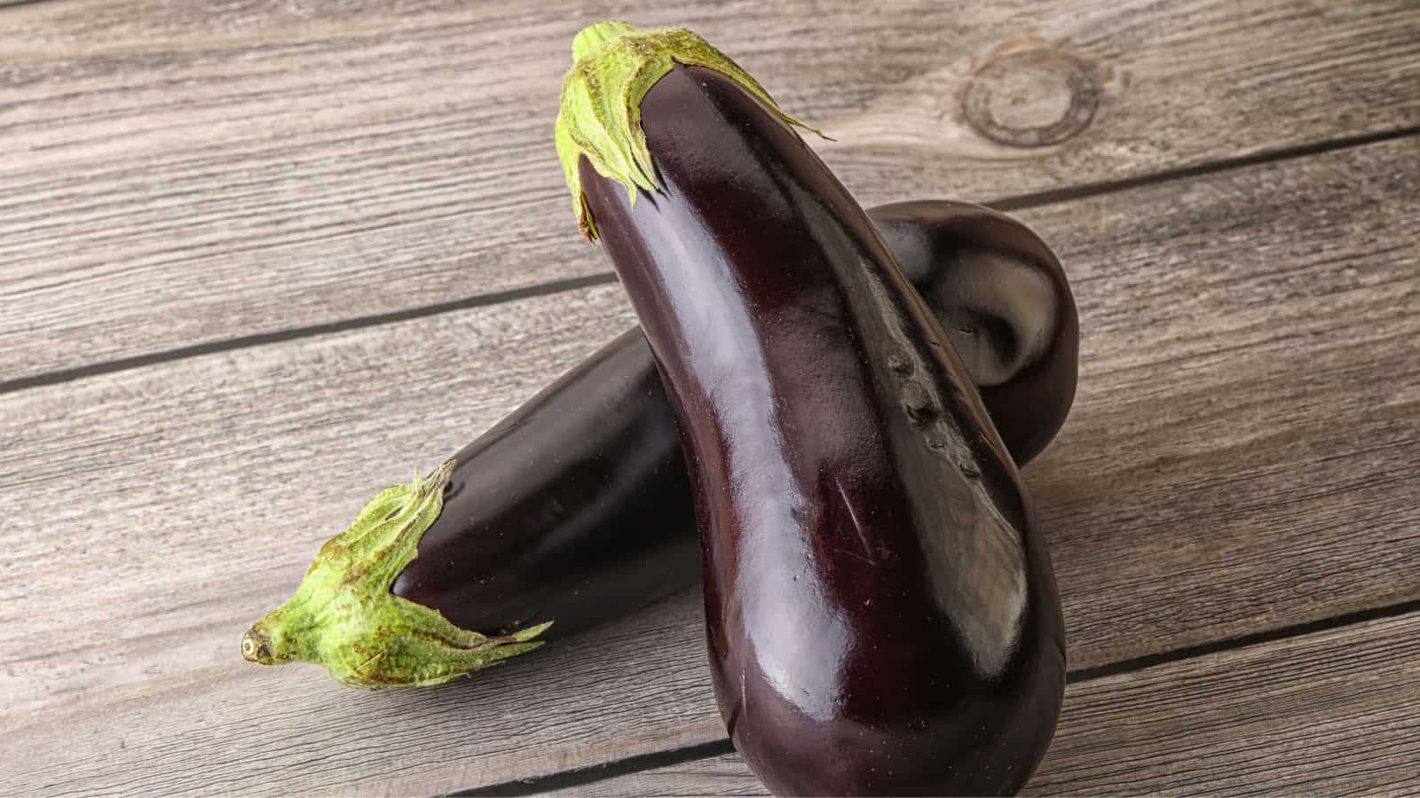 Two purple eggplants with green stems rest on a wooden surface. The eggplants are positioned diagonally, with one slightly overlapping the other. The wood features visible grain patterns and a circular knot near the top.
