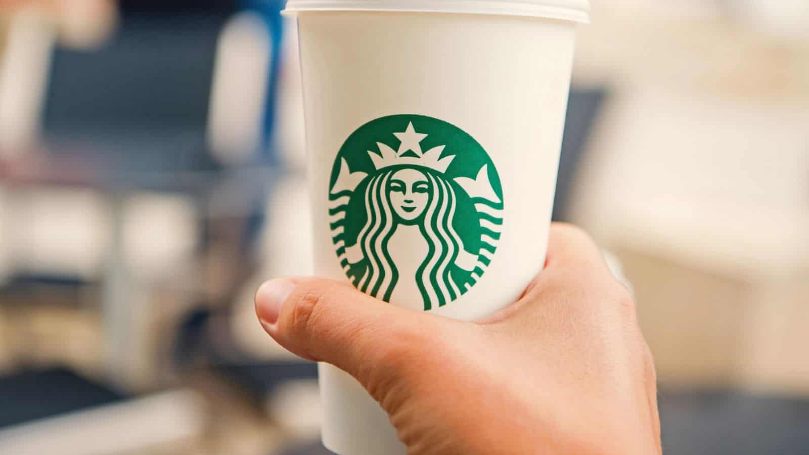 A hand is holding a white Starbucks cup with the green Starbucks logo on it. The background is blurred, showing indistinct indoor seating and tables.
