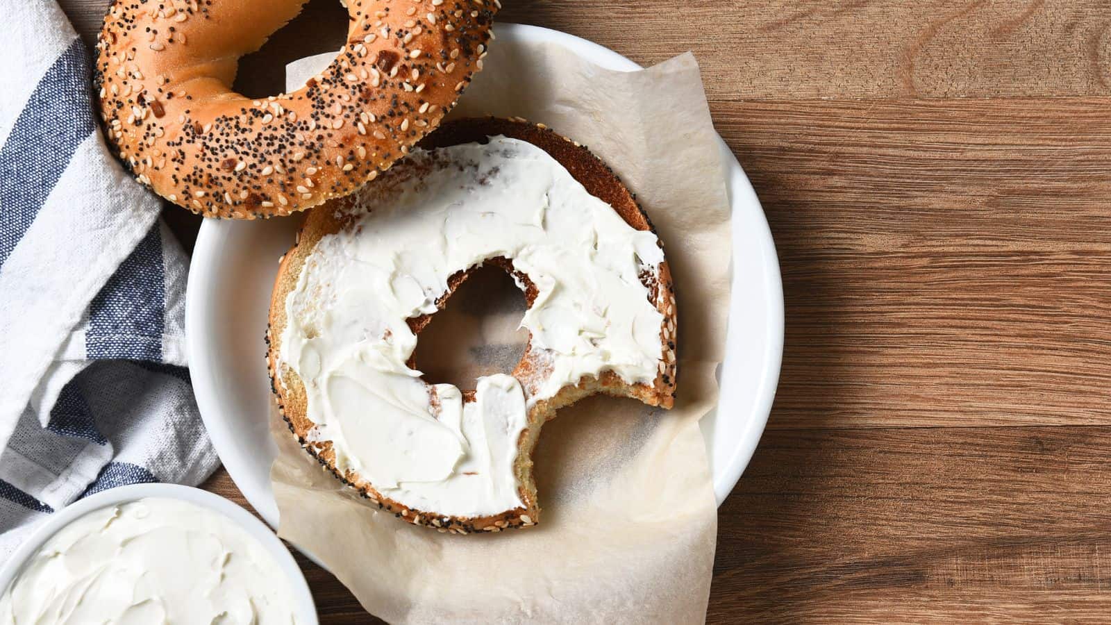 A partially eaten bagel with cream cheese sits on a white plate lined with parchment paper. Another whole bagel is nearby. A bowl of cream cheese and a striped cloth are next to the plate on a wooden surface.