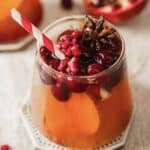 A glass filled with an orange-colored beverage, topped with cranberries, pomegranate seeds, and a star anise. A red and white striped straw is in the drink. An apple slice is visible inside. A pomegranate is partially visible in the background.