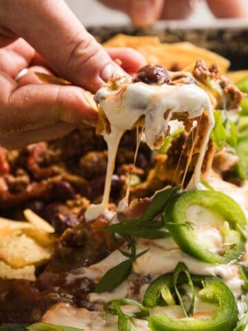A hand is lifting a nacho chip topped with melted cheese, ground meat, sliced jalapeños, and sour cream. The chip is part of a tray containing a larger serving of loaded nachos.
