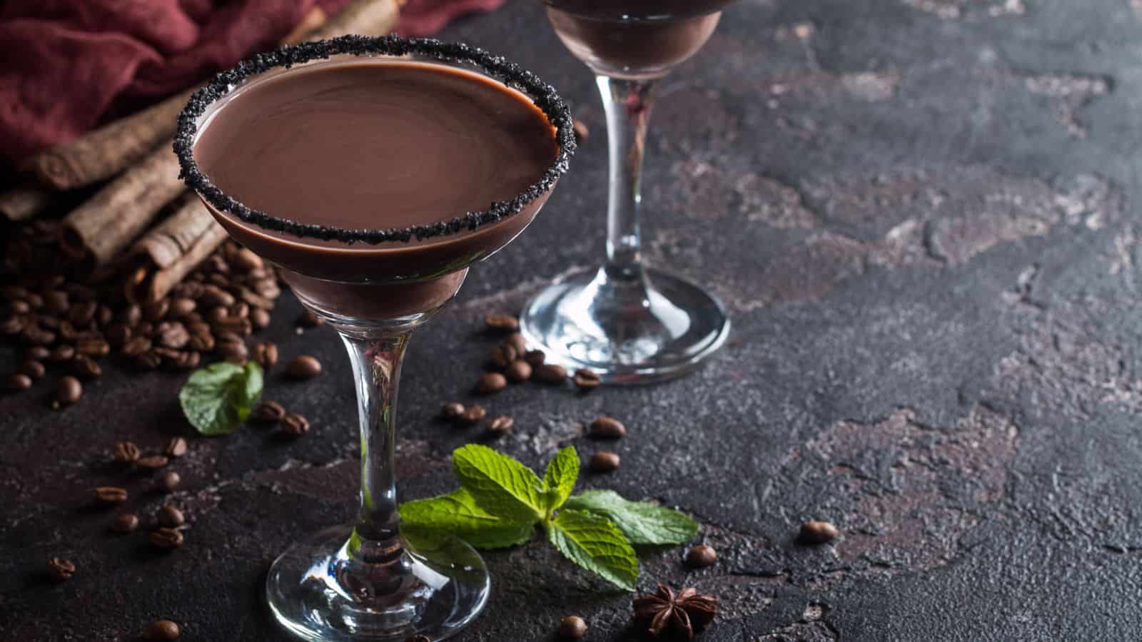 Two glasses filled with a chocolate cocktail sit on a textured dark surface. The rims are decorated with black sugar. Coffee beans, star anise, mint leaves, and cinnamon sticks are scattered around, adding to the presentation.