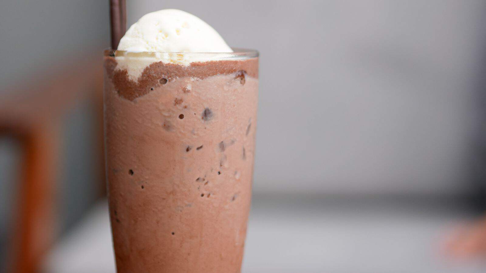 A tall glass of chocolate milkshake topped with a scoop of vanilla ice cream. The milkshake contains visible chocolate bits and is served with a straw. The background is blurred, focusing attention on the drink.