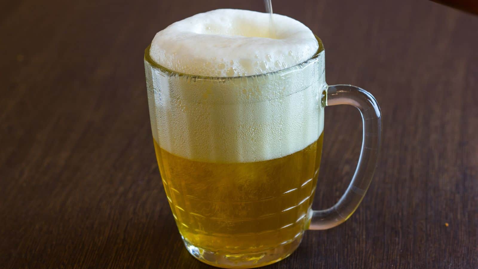 A glass mug filled with light golden beer sits on a dark wooden surface. The beer has a thick, frothy white head rising above the rim of the mug.