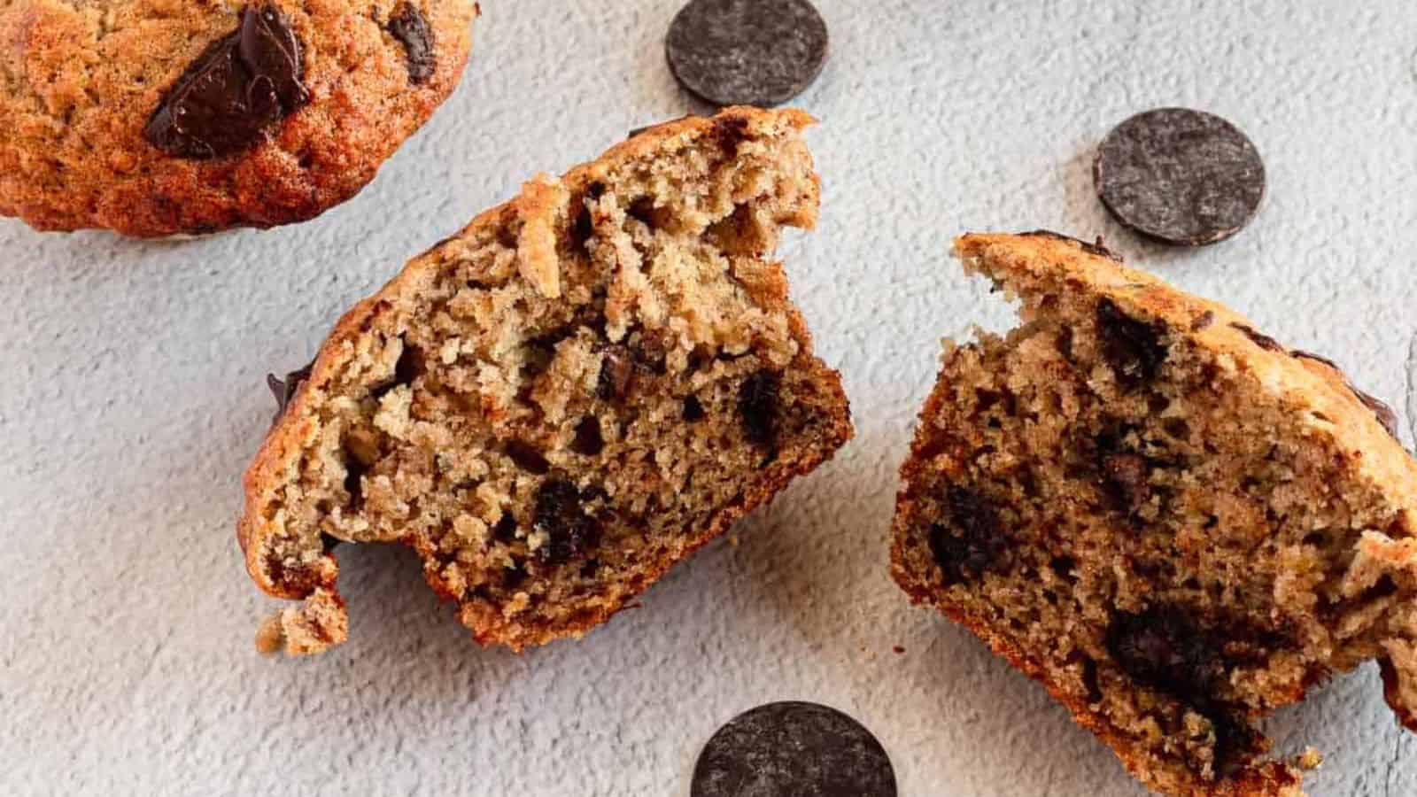 A halved chocolate chip muffin on a textured surface, showcasing its moist interior and chocolate pieces. Several chocolate wafers are scattered around, and another whole muffin is partially visible in the background.