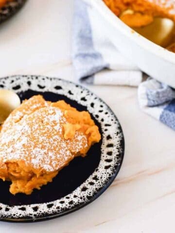 A slice of dessert topped with powdered sugar on a black and white plate, accompanied by a gold spoon. In the background, a white dish with more of the dessert and a blue and white cloth is visible. Another smaller plate with dessert is partially seen.