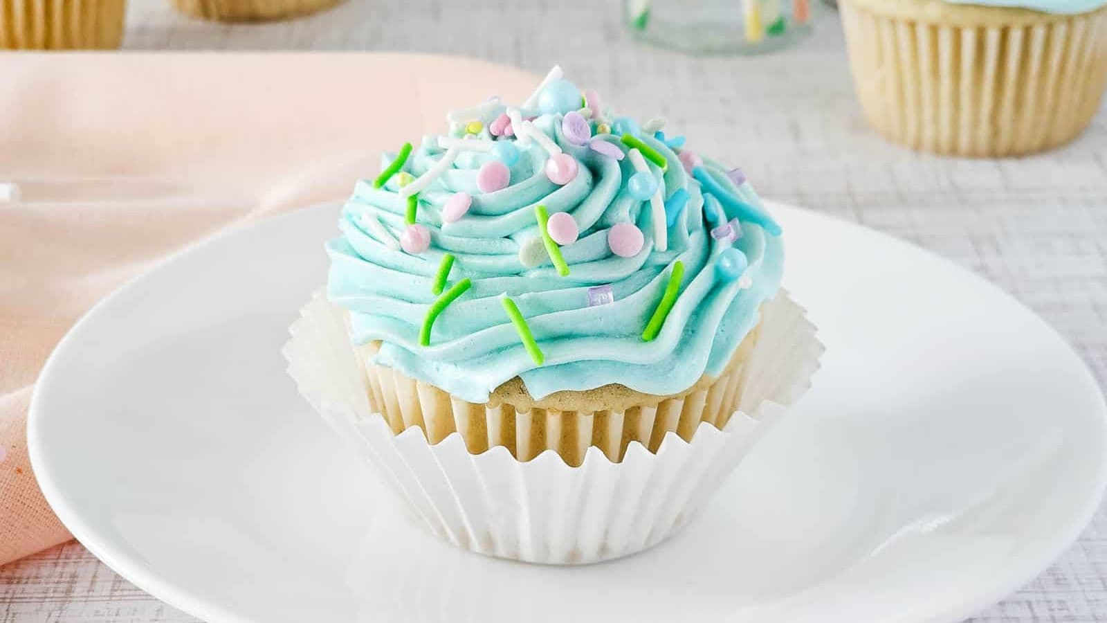 A vanilla cupcake with light blue frosting sits on a white plate. The frosting is topped with pastel sprinkles and green icing. The cupcake is in a white paper liner. A light pink cloth is partially visible in the background.