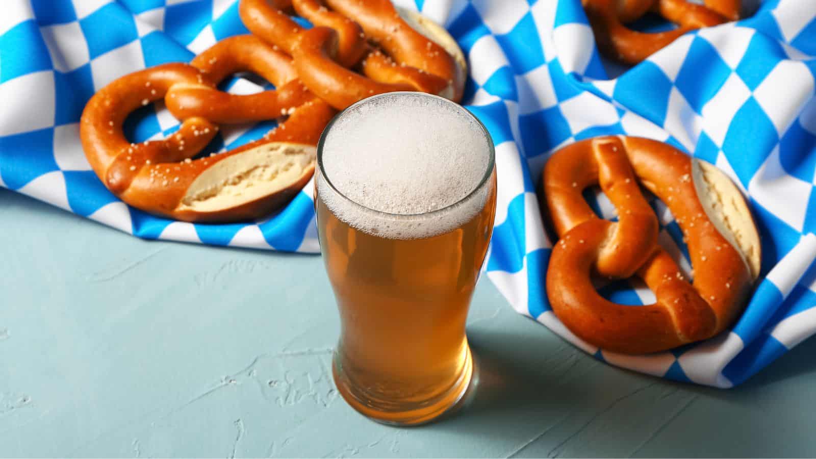 A glass of beer with a frothy head is placed on a light blue surface. Surrounding the glass are several pretzels on a blue and white checkered cloth. The pretzels have a golden-brown crust and are sprinkled with salt.