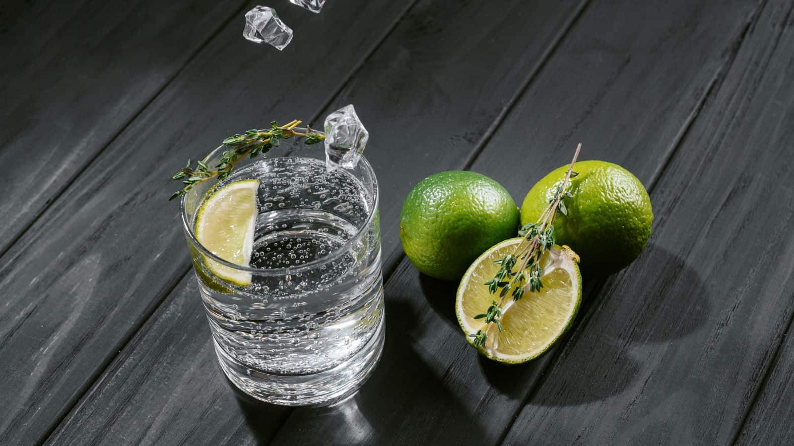 A glass of sparkling water with a lime slice and thyme sprig sits on a dark wooden surface. Ice cubes are being dropped into the glass. Two whole limes and a lime half garnished with a thyme sprig are beside the glass.