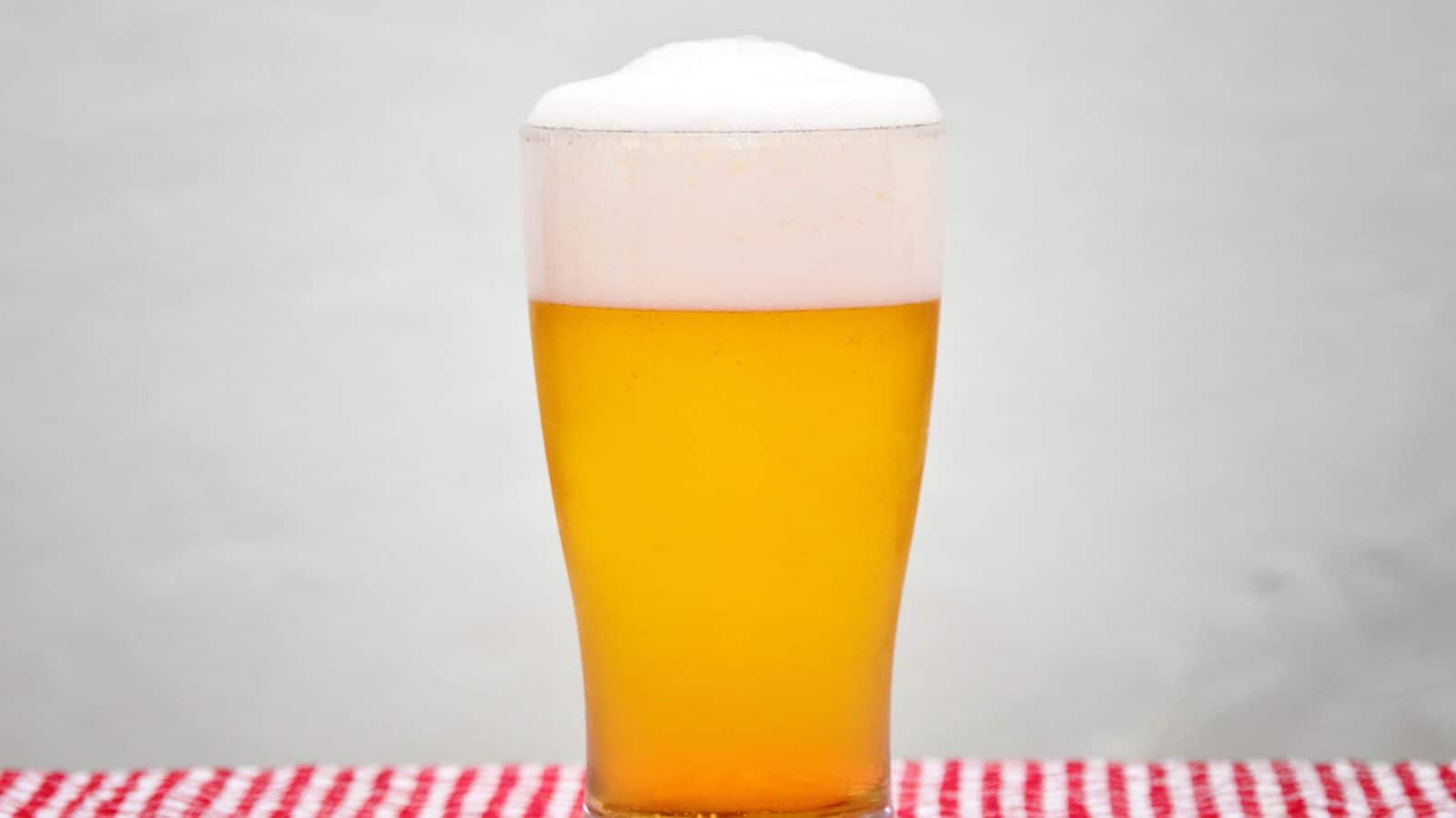 A tall glass of light beer, topped with a frothy white foam head, is placed on a red and white checkered surface against a plain background.