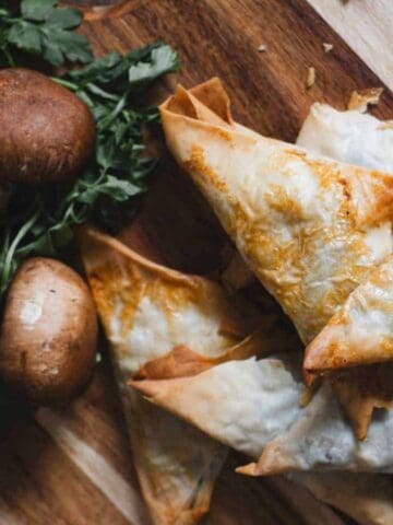 A wooden board displays cooked triangular pastries accompanied by whole mushrooms, shallots, and fresh herbs. The pastries have a golden, flaky appearance, while the mushrooms and shallots provide a rustic background.