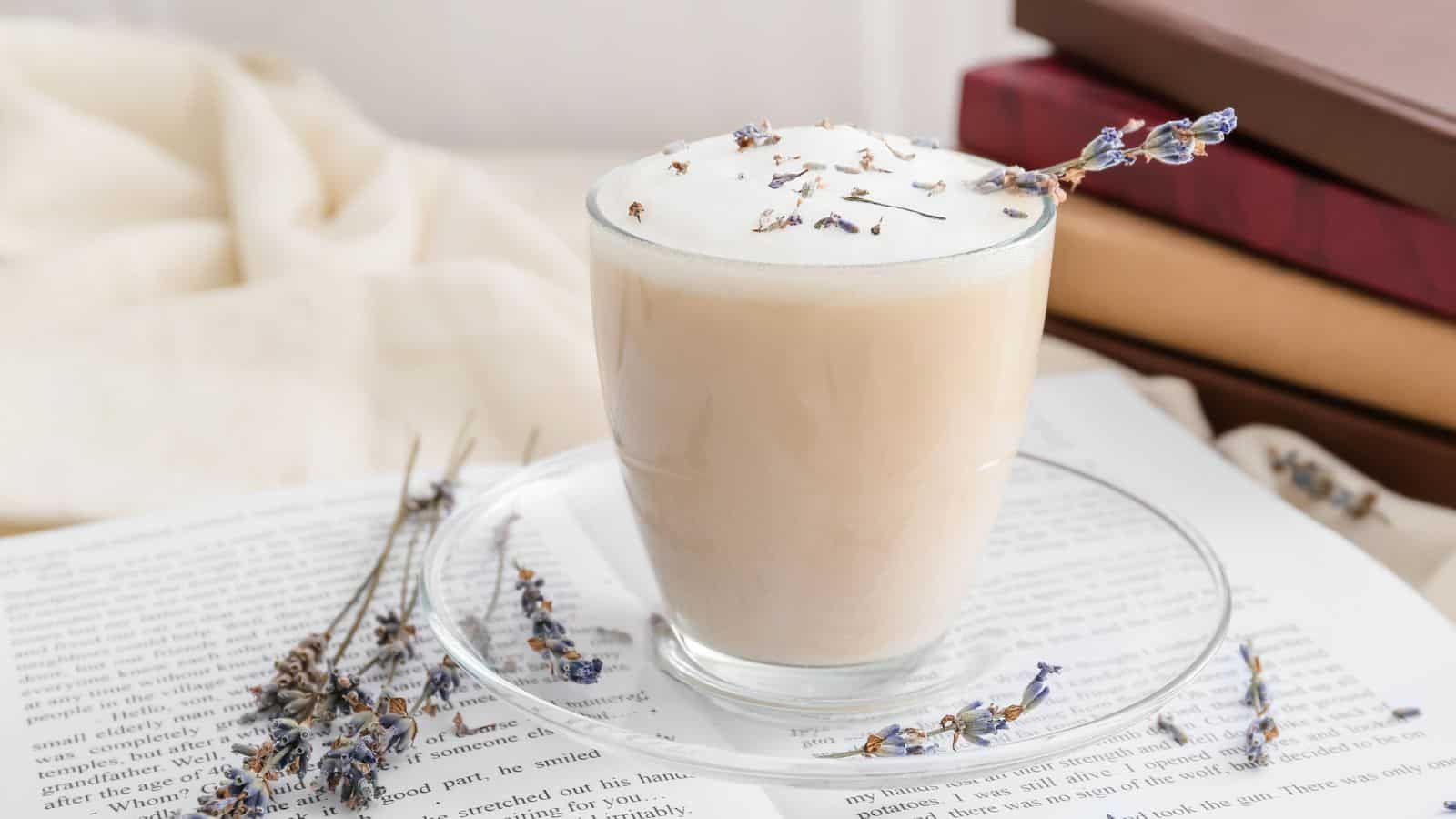 A glass of frothy milk topped with lavender flowers sits on a clear saucer. Dried lavender is scattered on an open book beneath. Books stacked in the background and a soft fabric enhance the cozy atmosphere.