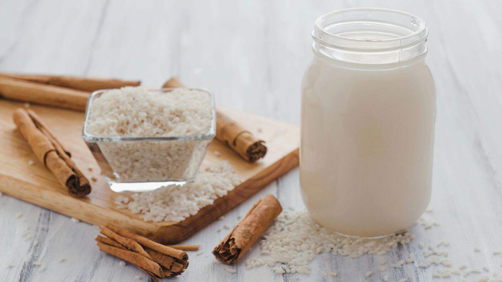 A jar of rice milk is placed on a light wooden surface. Next to it is a small glass bowl filled with uncooked rice, surrounded by scattered rice grains and cinnamon sticks on a cutting board. Additional cinnamon sticks are nearby.
