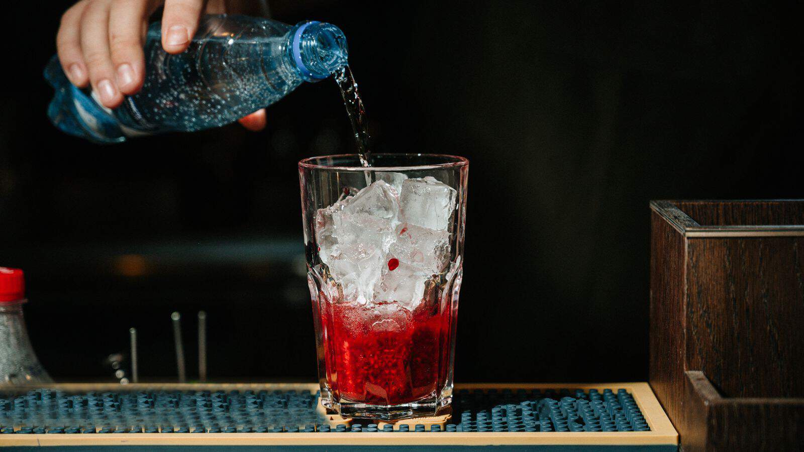 A hand pours clear liquid from a blue plastic bottle into a glass filled with ice cubes and red liquid, likely a cocktail, on a bar counter. A bar mat and a small part of a wooden structure are visible in the background.