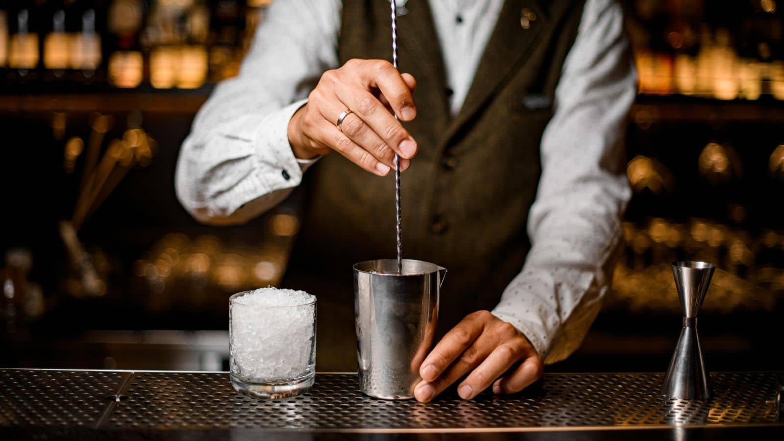 Bartender stirring a drink in a metal shaker using a long bar spoon. A glass filled with crushed ice is on the left, and a jigger is on the right. The bartender wears a white shirt and a dark vest. Bottles are blurred in the background.