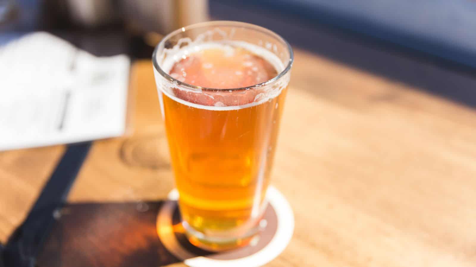 A clear glass filled with amber-colored beer sits on a wooden table. The glass is on a round coaster, and the drink has a light foam on top. Sunlight casts a shadow on the table. There are blurred objects in the background.